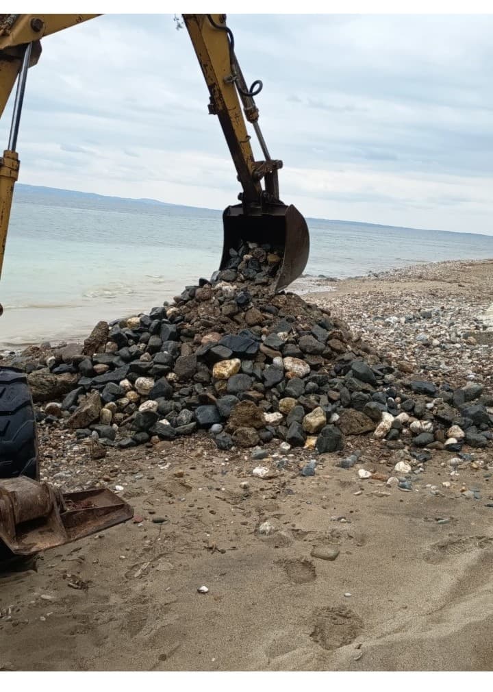 Εκσκαφέας μαζεύει πέτρες από την ακτή, με τη θάλασσα πίσω